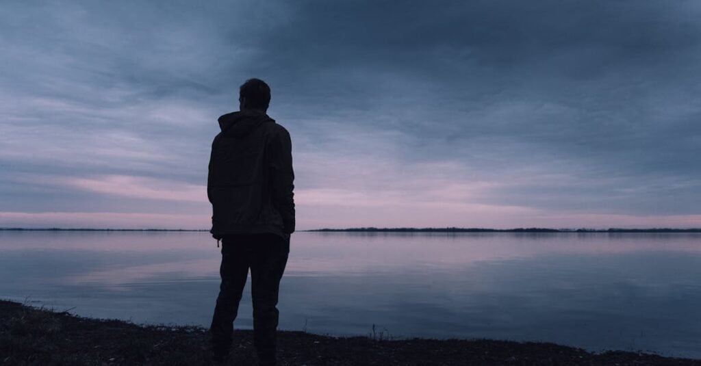 A solitary silhouette of a man in a jacket gazing at a lake during a peaceful sunset, creating a serene atmosphere.